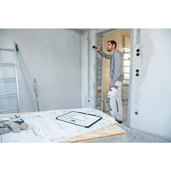 Bosch Professional Laserentfernungsmesser GLM 50-27 CG - Anzahl Akkus: 1 Akku, Messbereich: 0,05 - 50 m - Ein Mann misst mit einem Laser-Entfernungsmesser einen Raum, während Baupläne und ein Tablet auf einem Tisch liegen.