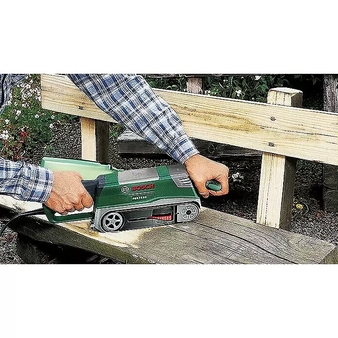 Bosch Lijadora de banda eléctrica de sobremesa PBS 75 AE - 750 W - Una persona utiliza una lijadora de banda Bosch verde y plateada (PBS 75 AC) para lijar un banco o valla de madera al aire libre.