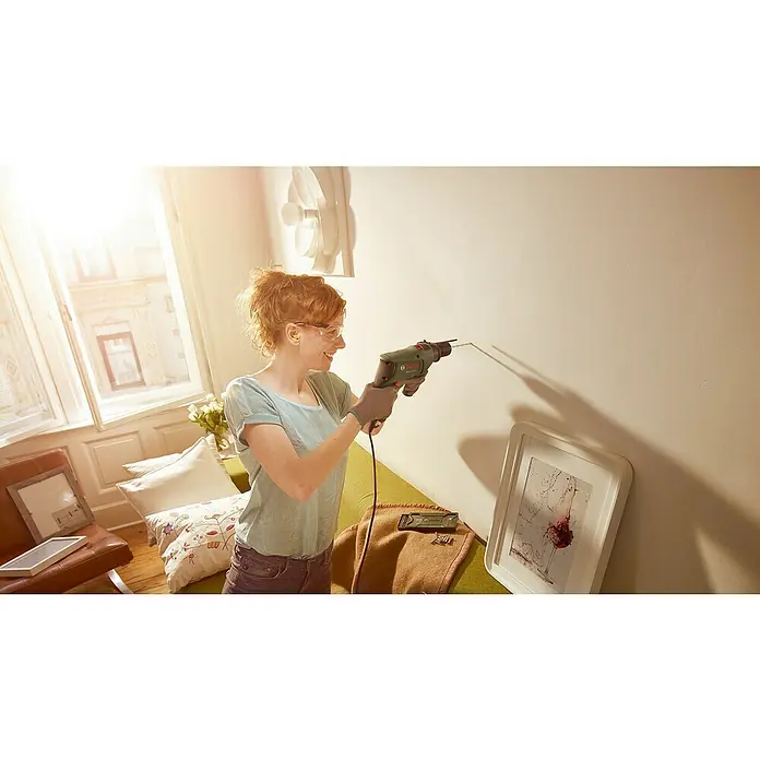 Bosch Schlagbohrmaschine EasyImpact 600 - 600 W, Leerlaufdrehzahl 1. Gang: 45.000 U/min - Eine Frau bohrt mit einer grünen Bosch-Bohrmaschine ein Loch in eine Wand, um ein Bild aufzuhängen. Sie trägt dabei eine Schutzbrille.