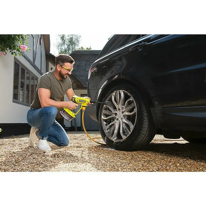 Ryobi Waschbürste RAC749 - Geeignet für: Felgen - Ein Mann kniet und benutzt einen gelben Ryobi-Akku-Reiniger, um ein Rad eines schwarzen Autos zu reinigen, mit einem Haus im Hintergrund.