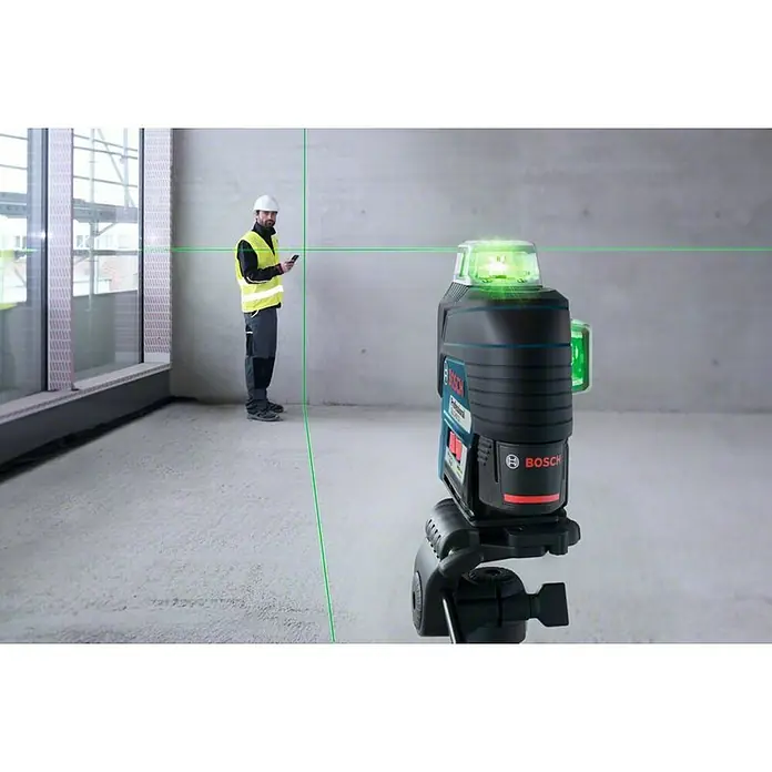 Bosch Professional Láser de línea GLL 3-80 CG - Zona de trabajo máx.: 30 m - El láser de líneas cruzadas Bosch proyecta líneas verdes sobre paredes de hormigón y el suelo mientras un trabajador de la construcción está cerca con un teléfono inteligente.