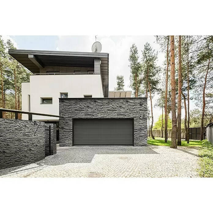 Stegu Verblendstein Mesada - 12 x 37 cm, Graphit, Steinoptik - Ein dunkelgraues Garagentor mit horizontalen Paneelen, integriert in eine moderne Hausfassade aus dunklem Stein, mit einer Kopfsteinpflasterauffahrt.