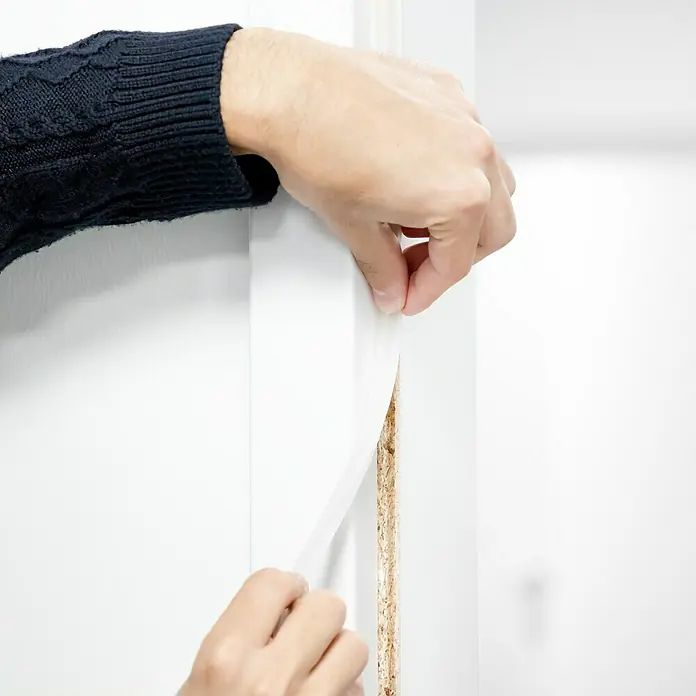 Primo Türdichtung Lignum 34 - Transparent, 5 m, Passend für: Nutbreite 3 – 4 mm - Eine Person, die eine weiße Folie von einer Holztür entfernt. Nahaufnahme der Hand, die die Folie abzieht und die Holzstruktur freilegt.