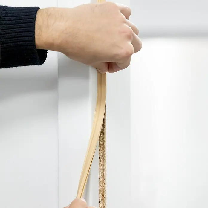 Primo Türdichtung Lignum 34 AC - Beige, 5 m, Passend für: Nutbreite 3 – 4 mm - Eine Person befestigt eine hellbraune Zierleiste an einer weißen Tür, um ihr Aussehen zu verbessern und einen sauberen Übergang zu schaffen.
