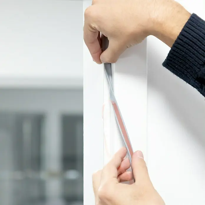 Primo Universaldichtung Densa 25 - Grau, 6 m, Passend für: Fenster, Türen, Möbel mit Spaltenbreite 2 – 5 mm - Jemand befestigt ein graues Dichtungsband mit Klebestreifen an einem weißen Türrahmen.