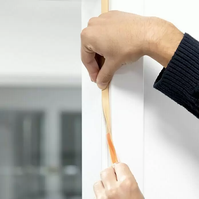 Primo Universaldichtung Densa 25 - Beige, 6 m, Passend für: Fenster, Türen, Möbel mit Spaltenbreite 2 – 5 mm - Hände bringen beiges Dichtungsband an einen weißen Rahmen an, wobei die Klebeschicht sichtbar wird.
