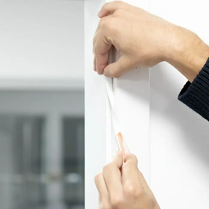 Primo Universaldichtung Densa 37 - Weiß, 6 m, Passend für: Fenster, Türen, Möbel mit Spaltenbreite 3 – 7 mm - Eine Person bringt ein weißes Dichtungsband an einem weißen Türrahmen an, um Zugluft zu verhindern.