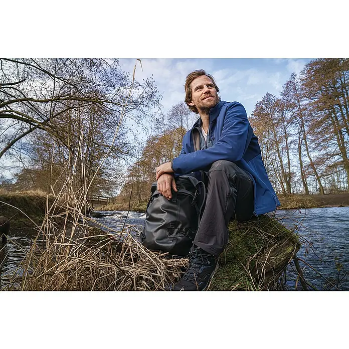 Tesa Ultra Power Reparaturgewebeband Extreme - Schwarz, 10 m x 50 mm - Ein Mann mit Rucksack sitzt an einem Flussufer, die blaue Jacke und der Rucksack sind gut sichtbar, während er die Natur genießt.