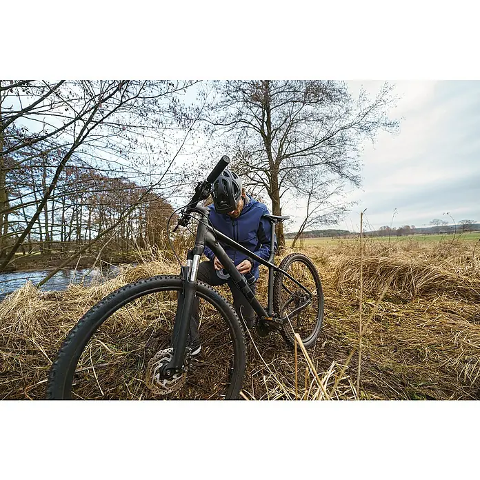 Tesa Ultra Power Reparaturgewebeband Extreme - Schwarz, 10 m x 50 mm - Ein Mann repariert sein schwarzes Cube Mountainbike in der Natur, vor einem Feld und Bäumen, mit Helm und blauer Jacke.