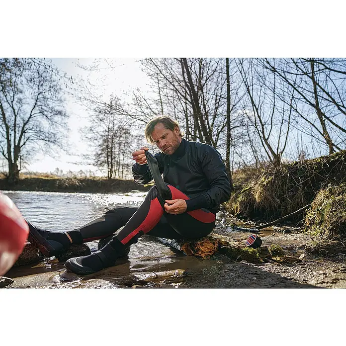 Tesa Ultra Power Unterwasser-Klebeband Under Water - Schwarz, 1,5 m x 50 mm - Ein Mann in einem Neoprenanzug sitzt am Flussufer und zieht einen schwarzen Neopren-Beinschutz an, um sich vor Kälte zu schützen.