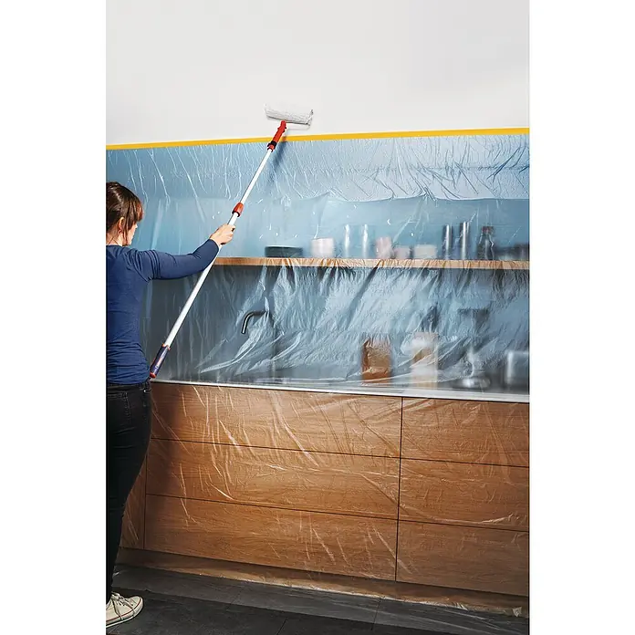 Tesa Abdeckfolie Easy Cover Perfect+ im Spender - L x B: 33 m x 140 cm - Eine Person benutzt eine Farbwalze mit Verlängerungsstange, um eine Wand zu streichen, während die Küche mit Folie abgedeckt ist.