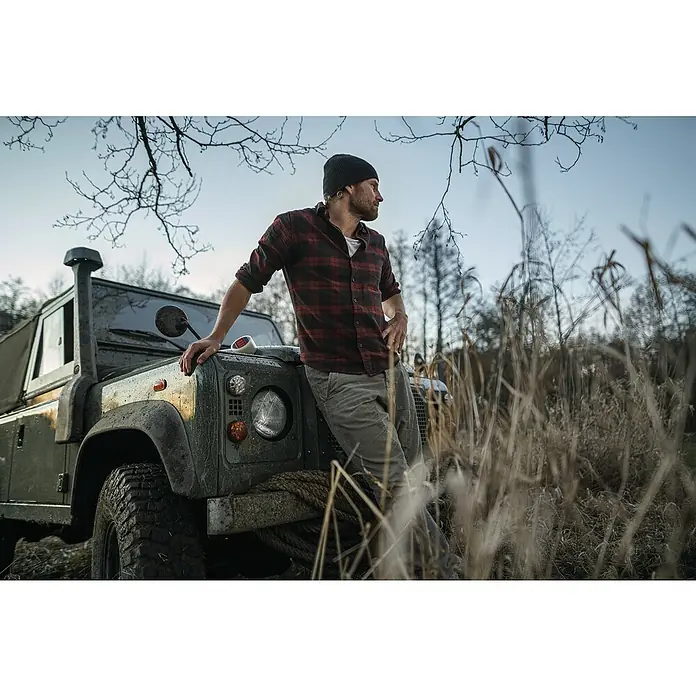 Tesa Ultra Power Reparaturklebeband Clear - Transparent, 10 m x 50 mm - Ein Mann in kariertem Hemd lehnt an einem schlammbedeckten Land Rover Defender im Gelände. Schwarze Mütze und derbe Reifen.