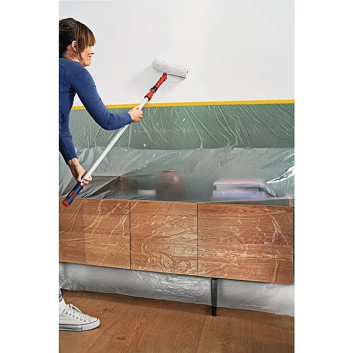 Tesa Abdeckfolie Easy Cover Perfect+ im Spender - L x B: 33 m x 140 cm - Frau streicht Wand mit Farbroller. Abdeckung schützt Möbel.