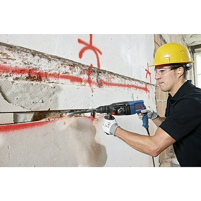 Bosch Professional Martillo combinado GBH 2-26 F - 830 W, 2,7 J, Alojamiento SDS-Plus - Un hombre usa un martillo perforador Bosch en una pared. Partículas de polvo vuelan en el aire.