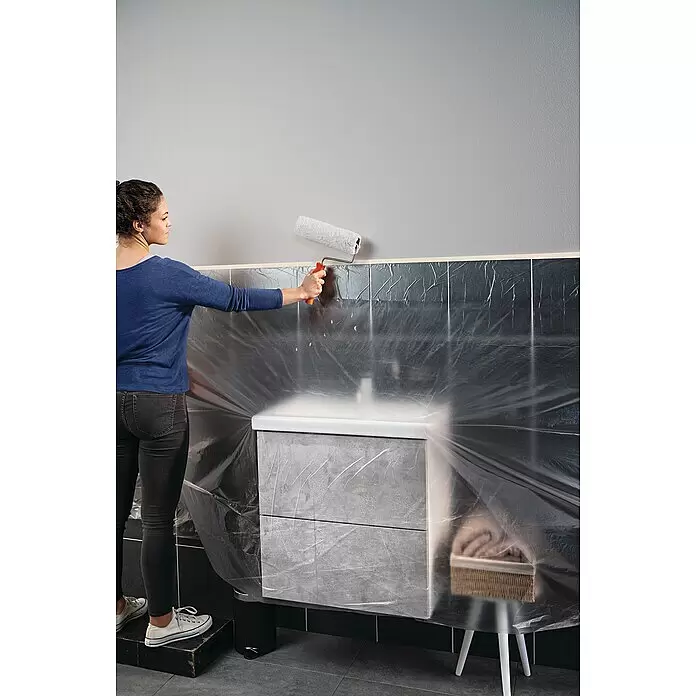Tesa Abdeckfolie Easy Cover Economy - L x B: 17 m x 260 cm - Frau streicht eine Wand mit einer Rolle. Badezimmermöbel mit Schutzfolie abgedeckt.