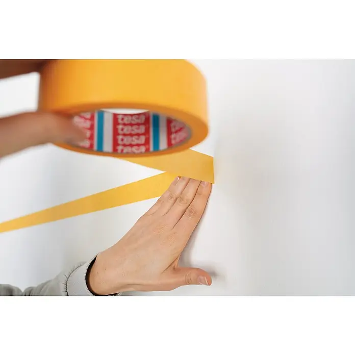 Tesa Malerband Perfect+ - 50 m x 30 mm - Gelbes Tesa-Band wird an einer weißen Wand angebracht. Die Rolle des Klebebands ist ebenfalls sichtbar, was die Verwendung des Produkts hervorhebt.