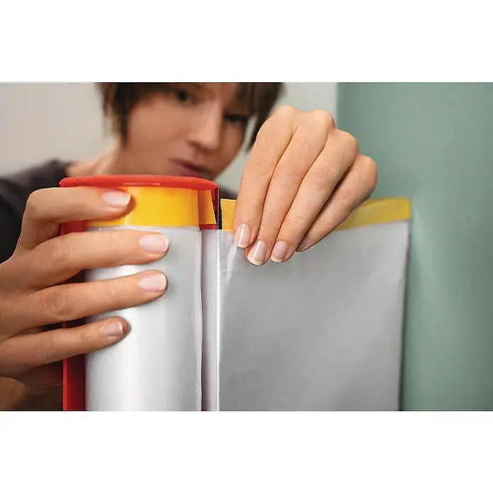 Tesa Abdeckfolie Easy Cover Perfect+ im Spender - L x B: 33 m x 140 cm - Nahaufnahme einer Person, die mit einem Abdeckband Abdeckfolie an einer Wand befestigt, um einen sauberen Farbauftrag zu gewährleisten.