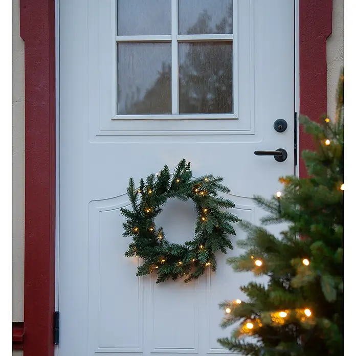 Eglo Figura LED navideña Guirnalda - 10 x 45 x 45 cm, Verde, LED - Una puerta festiva adornada con una corona navideña verde que brilla con luces cálidas y un pequeño árbol de Navidad colocado junto a la puerta.