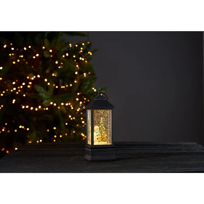Eglo Luz LED navideña Farolillo - 8,5 x 8,5 x 22 cm, Negro, LED - Imagen evocadora de una linterna negra con función de bola de nieve que muestra una escena invernal con un muñeco de nieve, colocada sobre una superficie de madera oscura.