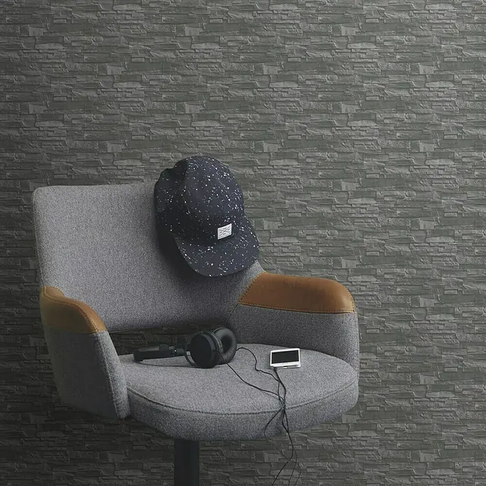 Rasch Vliestapete Steinoptik - Grau/Silber/Schwarz, 3D-Optik, 10,4 x 0,53 m - Nahaufnahme eines stilvollen grauen Stuhls mit Accessoires wie Kopfhörern, Mütze und MP3-Player vor einer Steinmauer.