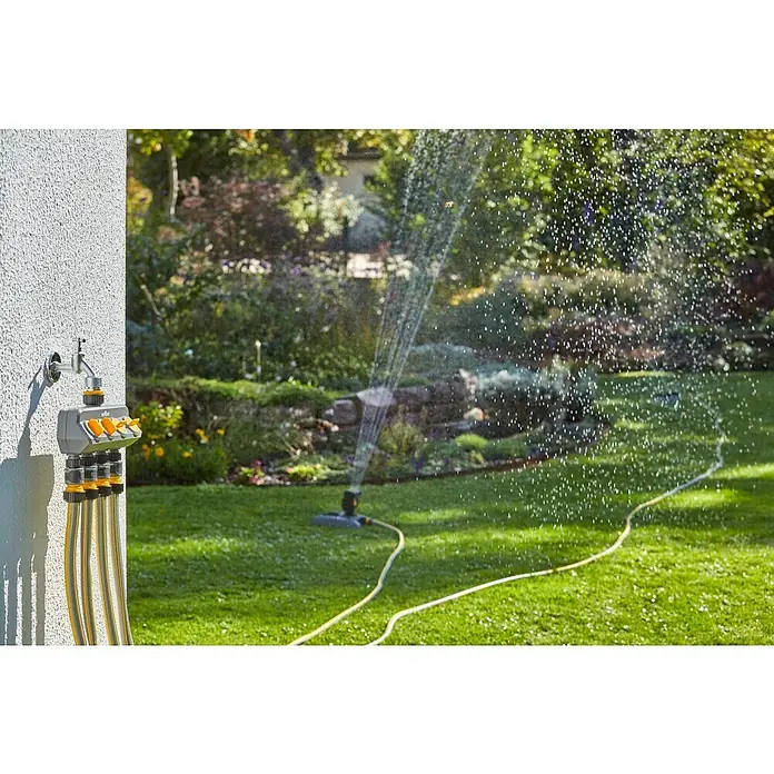 Neptun 4-Wege-Verteiler - Gewindegröße Wasserhahn: 33,3 mm (1″), 26,5 mm (¾″), 21 mm (½″) - Eine Mehrfachwasserhahn-Vorrichtung an einer Wand, mit gelben Schläuchen angeschlossen, während ein Rasensprenger im Garten läuft.