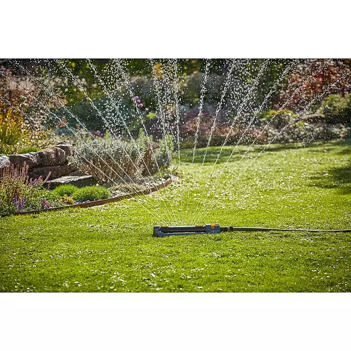 Neptun Viereckregner - Max. Regnerfläche: 460 m² - Ein grauer und oranger Oszillationsregner bewässert einen üppig grünen Rasen und Gartenpflanzen mit sichtbaren Wasserstrahlen.