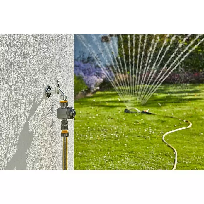 Neptun Bewässerungsuhr - Bewässerungsdauer: 5 min - 120 min - Ein silberner Wasserhahn an der Wand, verbunden mit einem grauen Bewässerungstimer und einem gelben Gartenschlauch, der einen Rasensprenger im grünen Garten versorgt.