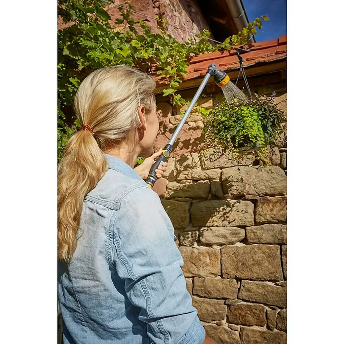 Neptun Gießstab - Länge: 76 cm, Aluminium/Kunststoff - Frau bewässert hängende Pflanzen mit einer gelben und grauen Bewässerungsdüse. Die Düse hat einen langen Griff für mehr Reichweite.