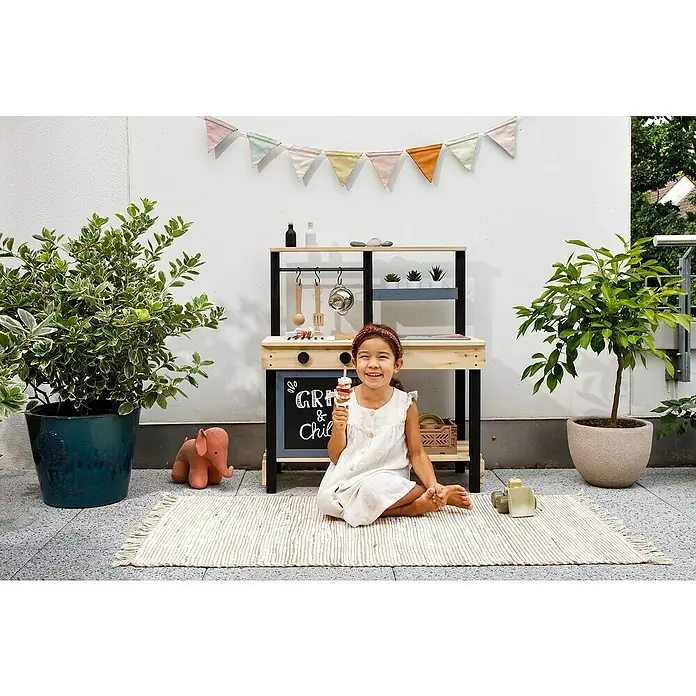 Muddy Buddy Matschküche Grill & Chill - 80 x 40 x 90 cm, Holz, Natur/Schwarz - Fröhliches Mädchen sitzt vor einer Spielküche mit Zubehör und hält ein Spielzeug-Eis am Stiel in der Hand.