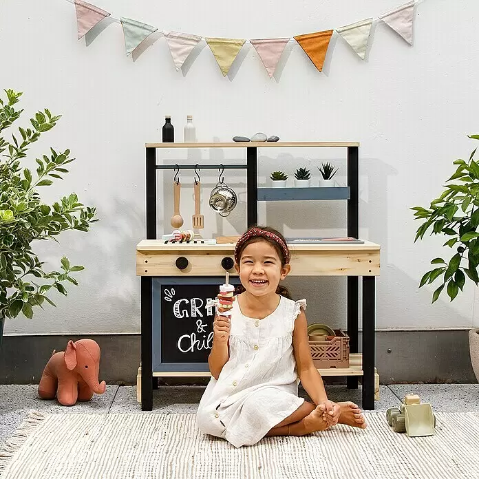Muddy Buddy Matschküche Grill & Chill - 80 x 40 x 90 cm, Holz, Natur/Schwarz - Lachendes Kind vor einer hölzernen Spielküche mit schwarzen und naturfarbenen Elementen, hält ein Spielzeug-Eis.