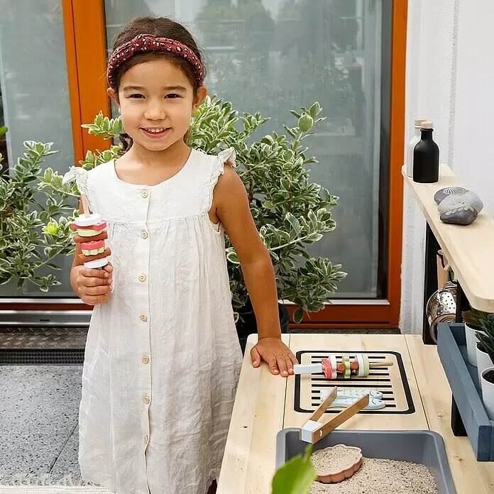 Muddy Buddy Matschküche Grill & Chill - 80 x 40 x 90 cm, Holz, Natur/Schwarz - Kind spielt mit einer Holzmatschküche, hält einen Spielzeug-Kebab. Das Set enthält Spielzeugessen, Zange und eine Sandwanne.