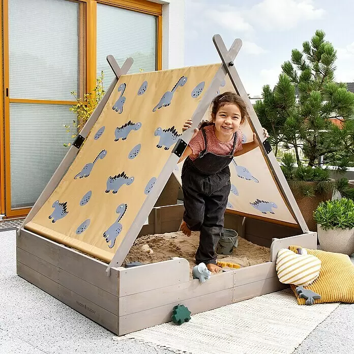 Muddy Buddy Sandkasten Dino Lover - 140 x 120 x 140 cm, Holz - Ein Kind spielt in einem grauen Holzsandkasten mit beigem Verdeck mit blauen Dinosaurier-Motiven.