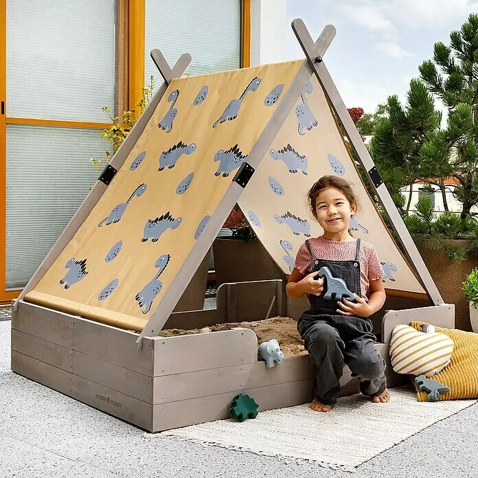 Muddy Buddy Sandkasten Dino Lover - 140 x 120 x 140 cm, Holz - Holzsandkasten mit Dinosaurier-Print-Vordach, in dem ein Kind mit Sandspielzeug sitzt. Der graue Holzkasten hat ein beiges Stoffdach.