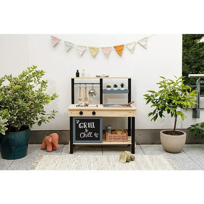 Muddy Buddy Matschküche Grill & Chill - 80 x 40 x 90 cm, Holz, Natur/Schwarz - Eine Outdoor-Spielküche aus Holz mit Grill und Zubehör steht auf einer Terrasse, umgeben von Pflanzen und Dekorationen.
