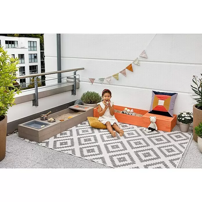 Muddy Buddy Sandkasten Flexy Style - 150 x 90 x 20 cm, Holz, Warmgrau/Lachs - Ein Mädchen spielt auf einem Balkon mit einer grau-orangen Holz-Sand- & Wasserspielstation auf einem weiß-grau gemusterten Outdoor-Teppich.