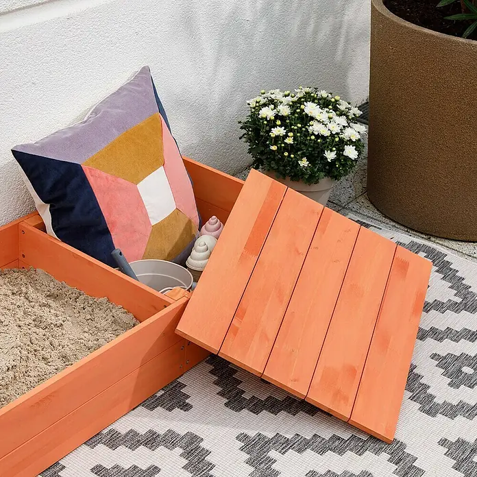 Muddy Buddy Sandkasten Flexy Style - 150 x 90 x 20 cm, Holz, Warmgrau/Lachs - Nahaufnahme einer orangefarbenen Kindersandkiste mit Abdeckung, gefüllt mit Sand, einem Kissen und Spielzeug auf einem gemusterten Teppich.