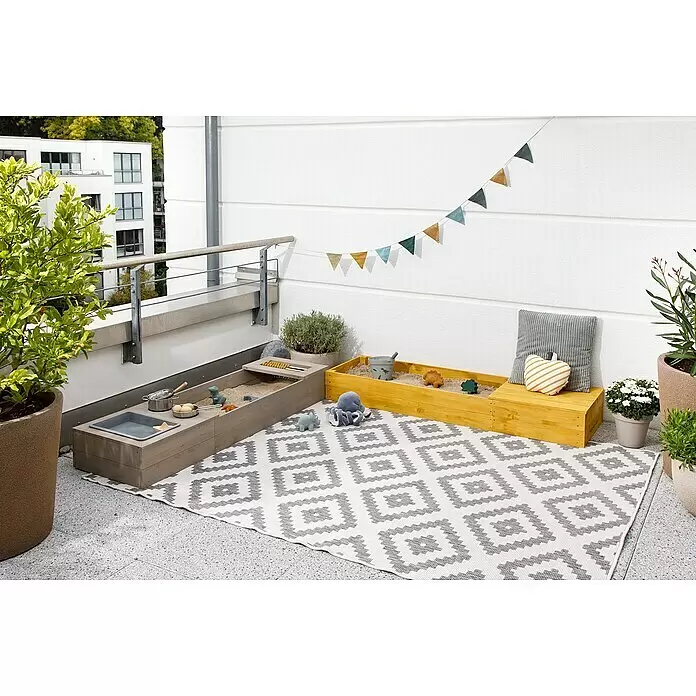Muddy Buddy Sandkasten Flexy Style - 150 x 90 x 20 cm, Holz, Warmgrau/Ocker - Zwei hölzerne Sandkästen, einer grau und einer gelb, auf einem gemusterten Outdoor-Teppich, gefüllt mit Sand und Spielzeug auf einem Balkon.