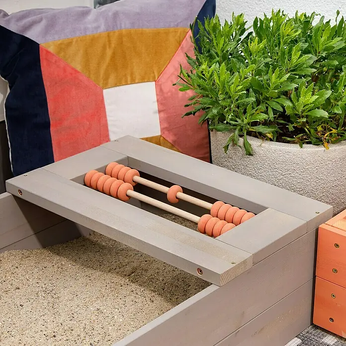 Muddy Buddy Sandkasten Flexy Style - 150 x 90 x 20 cm, Holz, Warmgrau/Lachs - Ein hellgrauer Sandkasten mit integriertem Abakus mit orangefarbenen Perlen und einem farbenfrohen Kissen im Hintergrund.
