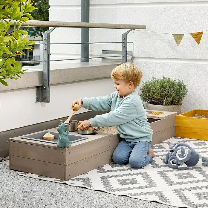 Muddy Buddy Sandkasten Flexy Style - 150 x 90 x 20 cm, Holz, Warmgrau/Ocker - Kind spielt mit einem Holzküchenspielset im Freien, das verschiedene Fächer und Spielutensilien umfasst.