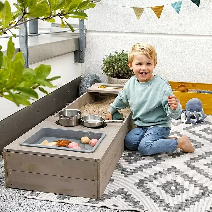 Muddy Buddy Sandkasten Flexy Style - 150 x 90 x 20 cm, Holz, Warmgrau/Ocker - Hölzerner Sand- und Wassertisch mit grauen Wannen, von einem Kind genutzt. Enthält Spielküchenzubehör für sensorisches Spiel.