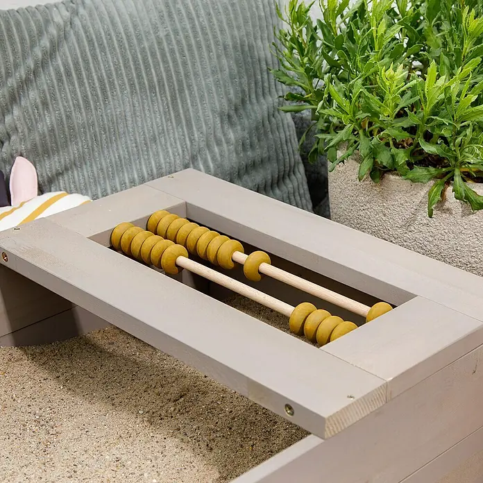 Muddy Buddy Sandkasten Flexy Style - 150 x 90 x 20 cm, Holz, Warmgrau/Ocker - Nahaufnahme eines Spieltisches mit Rechenrahmenaufsatz, darunter Sand. Ein dekoratives Kissen und eine Pflanze vervollständigen die Szene.