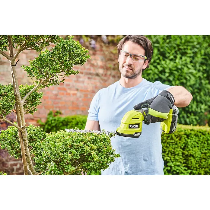 Ryobi ONE+ Akku-Gras- & Strauchschere RY18GSA-0 - 18 V, Li-Ionen, Ohne Akku, Schwertbreite: 12 cm - Ein Mann, der mit einer Ryobi Akku-Heckenschere einen Busch schneidet, mit Schutzbrille und Handschuhen für die Sicherheit.