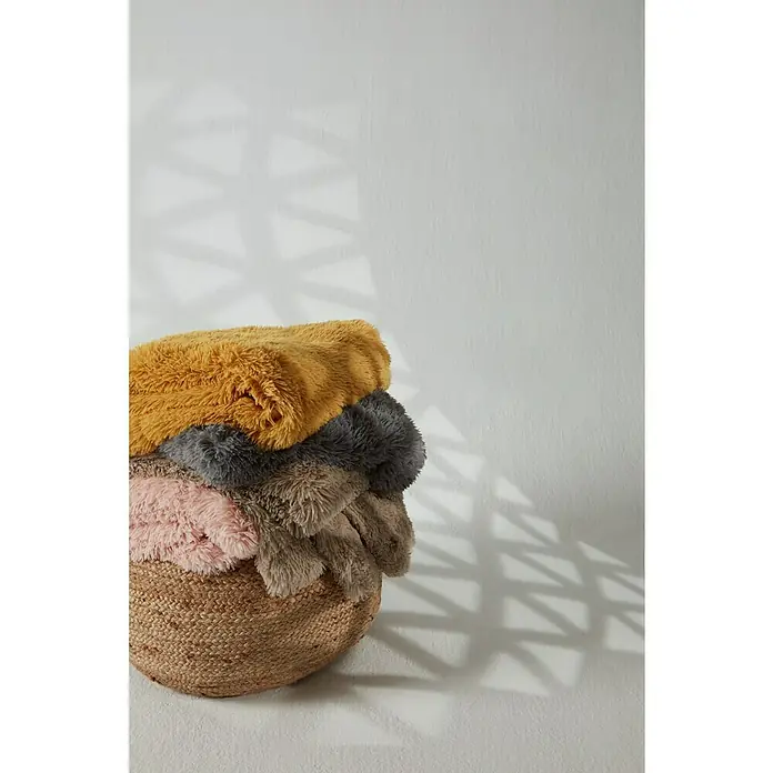 Manta para sofá Luxury - Multicolor - Una canasta llena de mantas coloridas y suaves, incluyendo amarillo mostaza, gris, rosa y marrón, sobre una pared blanca.