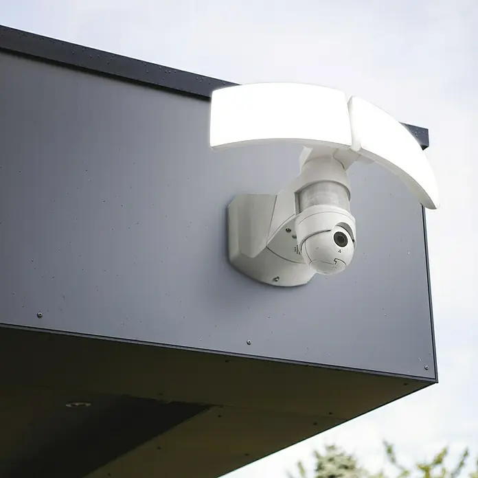 Lutec Proyector LED Libra doble con sensor y cámara - 38 W, L x An x Al: 227 x 309 x 202 mm, Blanco, Blanco frío - Primer plano de una cámara de seguridad blanca con focos en una pared gris, combinando vigilancia e iluminación.