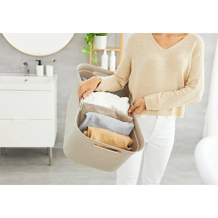 Rotho Wäschekorb Brisen - L x B x H: 59,6 x 39,6 x 23,2 cm, Kunststoff, Cappuccino - Frau hält einen Wäschekorb mit gefalteter Wäsche im Badezimmer. Der Korb ist beige und die Wäsche ist farblich sortiert.