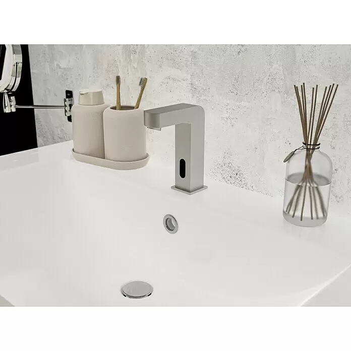 Camargue Kaltwasser-Standventil Sensor - Chrom, Glänzend - Modernes weißes Waschbecken mit automatischem grauen Wasserhahn, beigen Accessoires und Diffusor vor strukturierter grauer Wand.