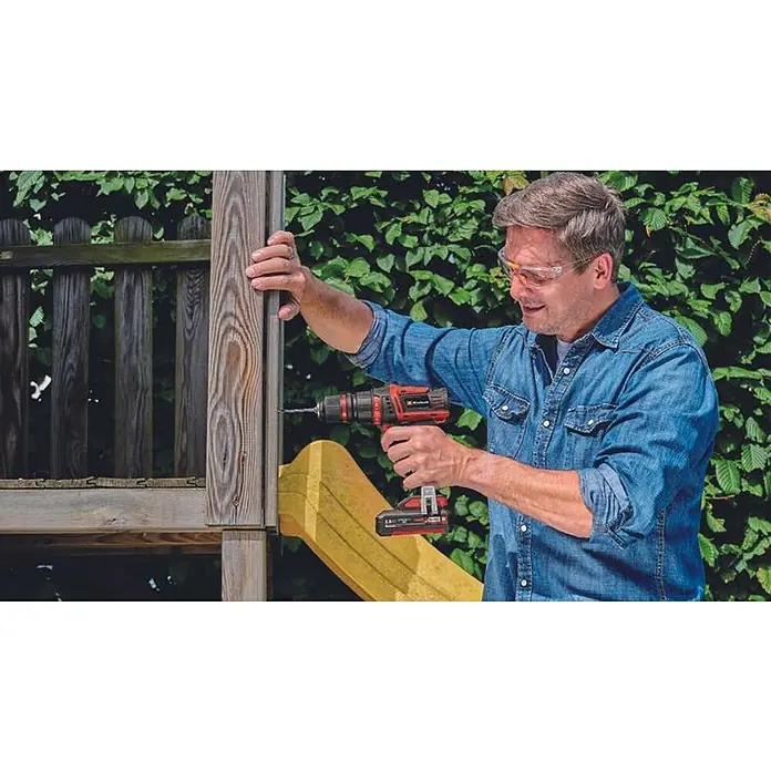 Einhell PXC 18V Taladro atornillador de batería TE-CD18/45 - 18 V, 2 baterías, 2 Ah, 45 Nm - Un hombre está usando un taladro inalámbrico Einhell para sujetar una pieza de madera, con follaje en el fondo. Lleva gafas de seguridad.