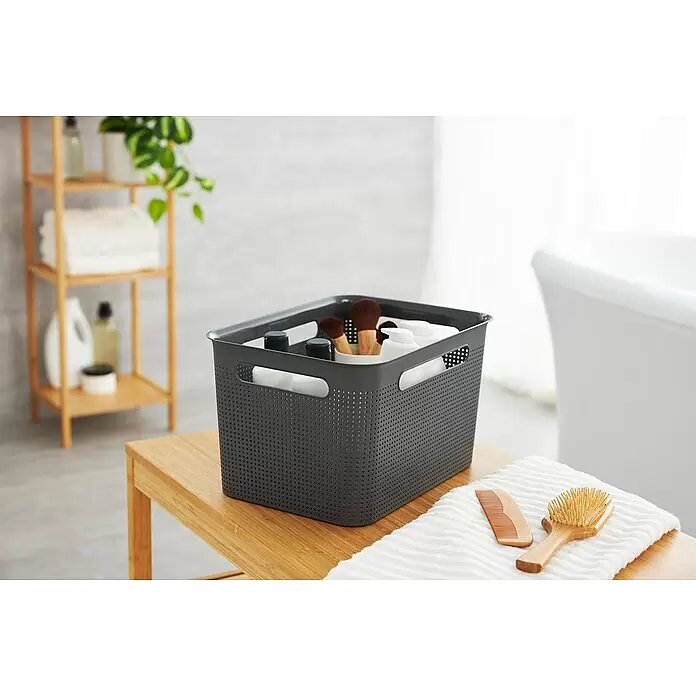 Rotho Aufbewahrungsbox Brisen - L x B x H: 36 x 26,2 x 21,1 cm, Kunststoff, Blau - Grauer, perforierter Aufbewahrungskorb auf einem Holzhocker im Bad, gefüllt mit Flaschen und Bürsten. Ein Kamm und eine Bürste liegen auf einem Handtuch.