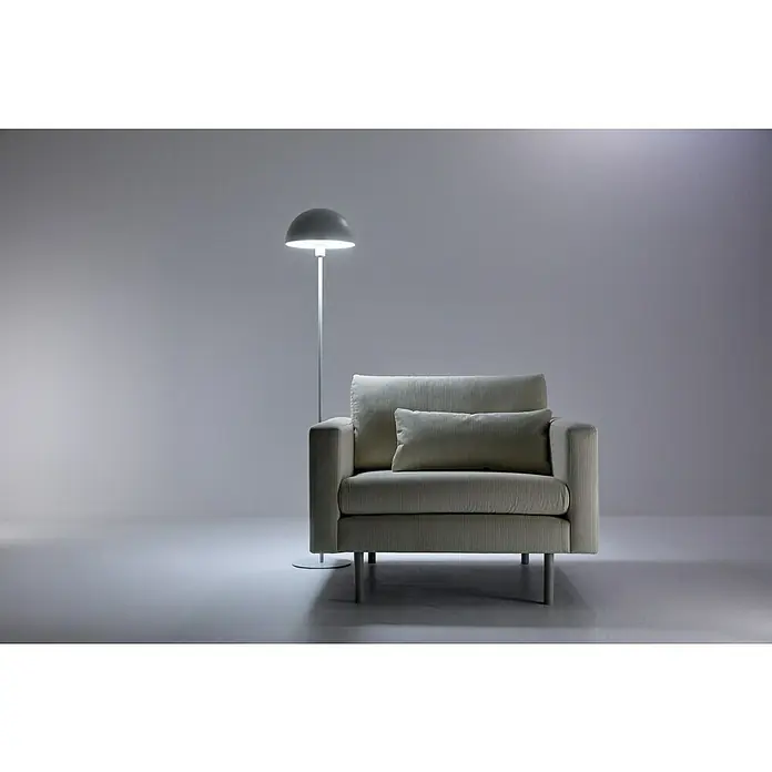 Philips Wiz Bombilla LED inteligente Full color - E27, RGBW, 806 lm, 8,5 W - Un elegante sillón con cojín y una lámpara de pie al lado, que proyecta una luz suave en la habitación. Mobiliario elegante para tu hogar.