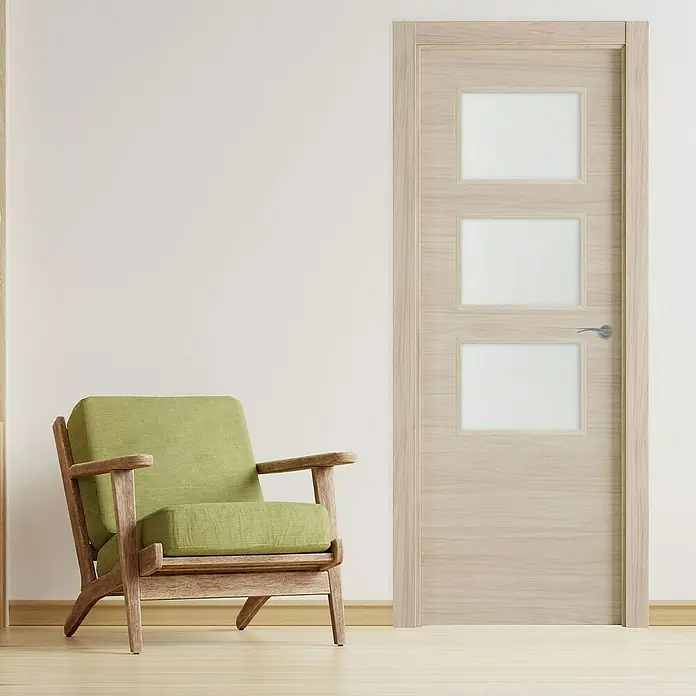 Pack puerta acristalada Arizona - 72,5 x 203 cm, Izquierda, Roble arena - Una puerta de madera clara con tres inserciones de vidrio, al lado de una silla verde en una habitación luminosa, diseño elegante.